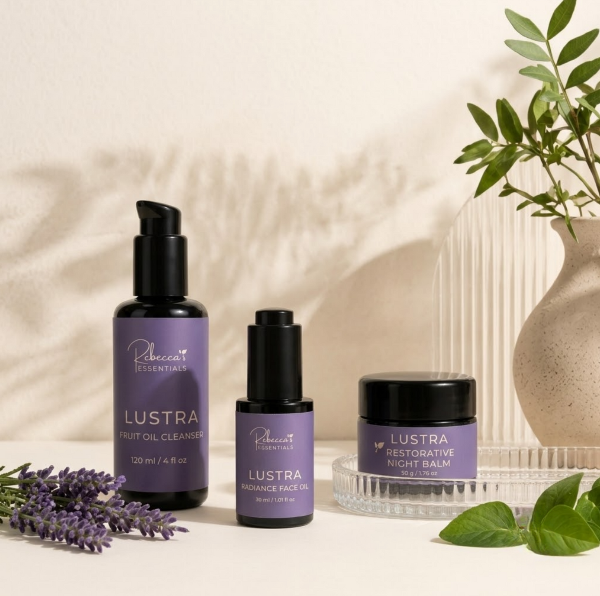 Three lavender-colored cosmetic products from Rebecca's Essentials including Lustra Fruit Oil Cleanser, Lustra Radiance Face Oil, and Lustra Restorative Night Balm, styled with lavender sprigs, green leaves, and a ceramic vase on a warm beige background.